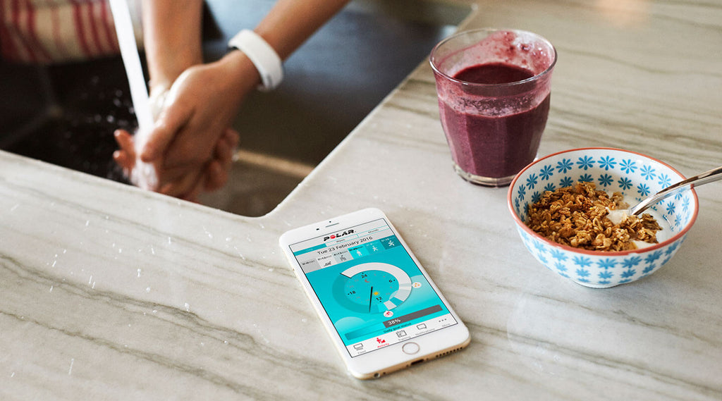 Polar app on mobile phone next to cereal bowl and a smoothie in a glass. Woman in background wearing Polar Loop 2 Activity Tracker