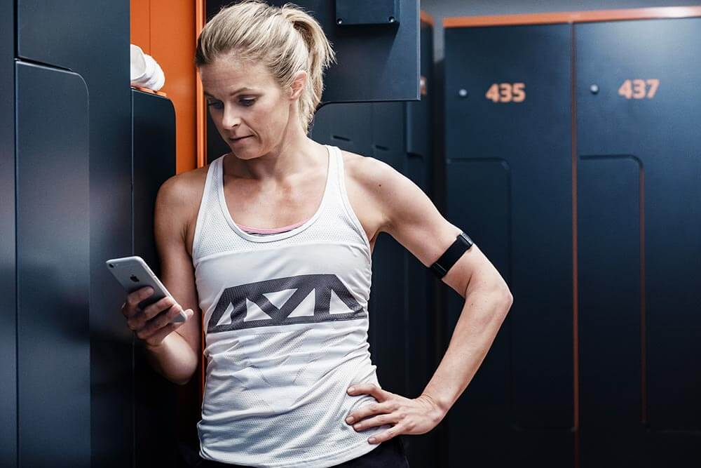 Woman wearing a Polar OH1 Arm Heart Rate Monitor whilst looking at the Polar app on her phone, standing next to the gym lockers
