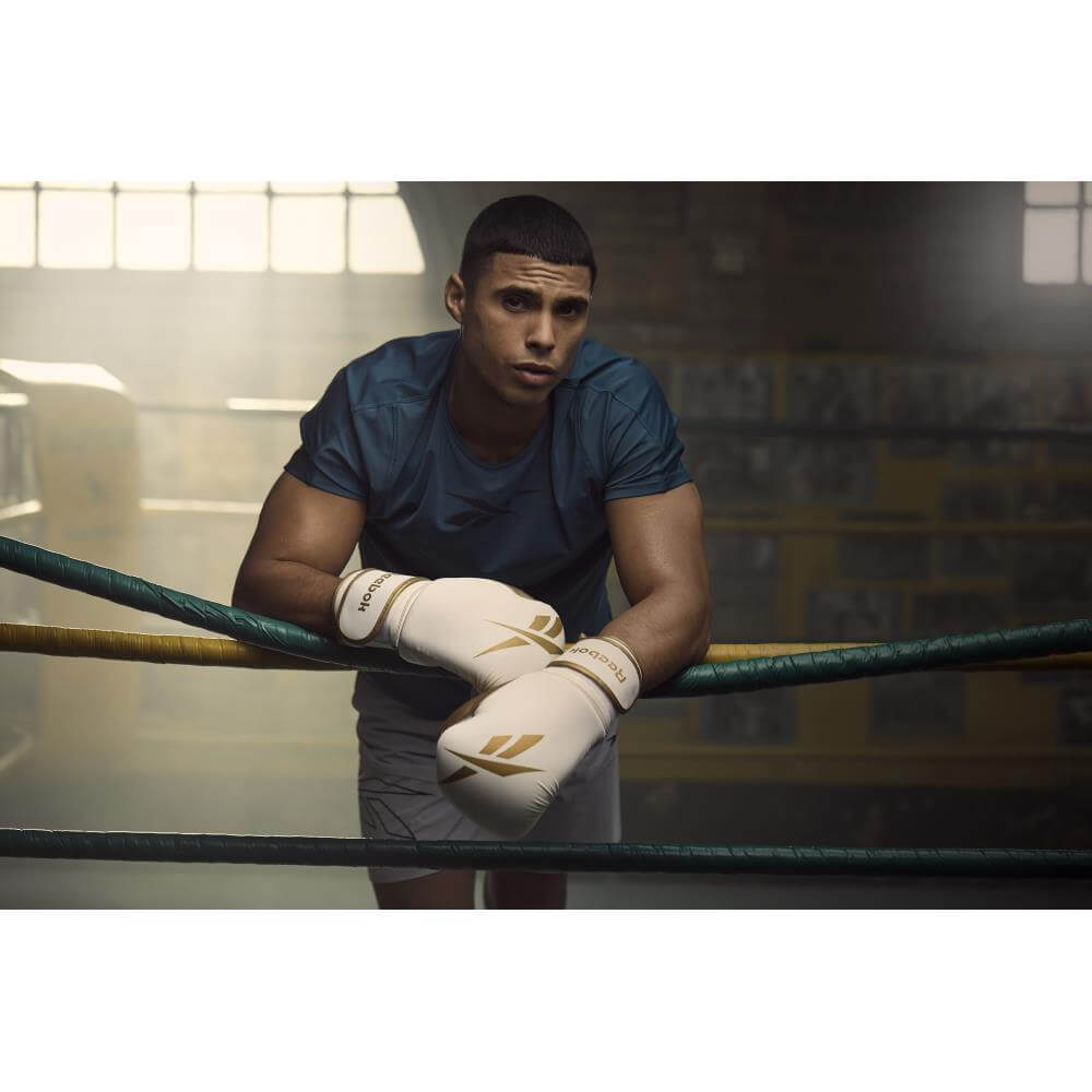 Man resting arms on boxing ring ropes whilst wearing Reebok Boxing Gloves - White and Gold